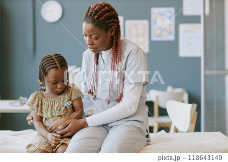 Medical professional in scrubs examining young child's arm in clinical setting. Child wearing dress while doctor attentively checks her health in bright, organized room 118643149