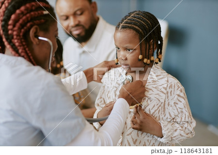 Child receiving medical examination with stethoscope by professional medical practitioner with other adults in background looking with concern Child receiving medical examination with stethoscope by professional medical practitioner with other adults in background looking with concern 118643181