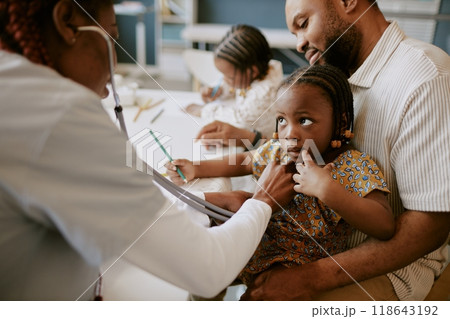 Child sitting on fathers lap holding his hand while doctor examining her. African American family in clinic setting with other children drawing in background 118643192