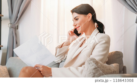 Business results. Positive woman. Mobile connection. Happy lady in elegant suit talking smartphone looking documents sitting sofa light room interior. Business results. Positive woman. Mobile connection. Happy lady in elegant suit talking smartphone looking documents sitting sofa light room interior. 118643308
