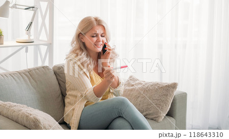 Desirable pregnancy. Happy woman. Mobile call. Cheerful lady looking pregnancy test talking smartphone sitting sofa light room interior. Desirable pregnancy. Happy woman. Mobile call. Cheerful lady looking pregnancy test talking smartphone sitting sofa light room interior. 118643310