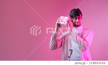 Romantic present. Valentine gift. Holiday surprise. Neon color light smiling handsome man in glasses showing box isolated on pink empty space background. Romantic present. Valentine gift. Holiday surprise. Neon color light smiling handsome man in glasses showing box isolated on pink empty space background. 118643416