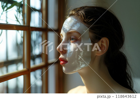 Close-up of a sheet mask on a face, emphasizing hydration and nourishment.A woman wearing a white mask is gazing out of a window 118643612