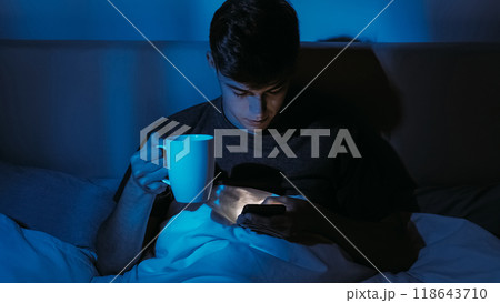 Relaxing atmosphere young man with coffee cup using phone in bed at night illuminated by the screen light. Relaxing atmosphere young man with coffee cup using phone in bed at night illuminated by the screen light. 118643710
