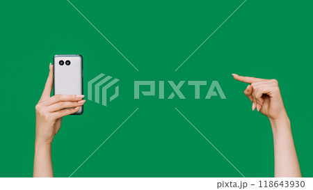 Mobile gadget. Online app. Modern technology. Female hand pointing finger showing smartphone back side isolated on green copy space advertising background. Mobile gadget. Online app. Modern technology. Female hand pointing finger showing smartphone back side isolated on green copy space advertising background. 118643930