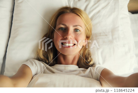 portrait of happy smiling young woman taking selfie with smartphone while lying in bed at home 118645224