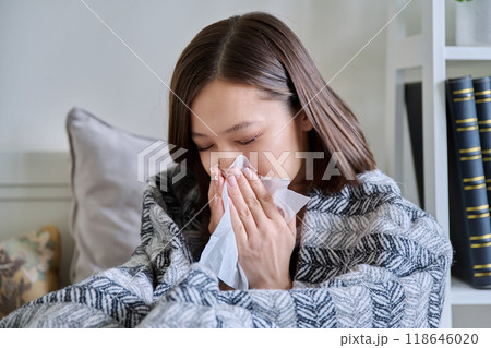 Young sick woman sitting under warm blanket sneezing coughing in handkerchief 118646020