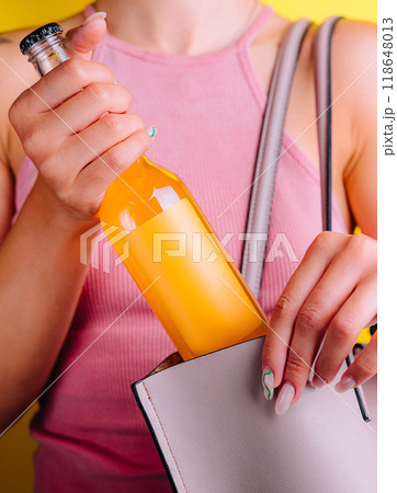 Woman hiding orange juice bottle in her bag on yellow background Woman hiding orange juice bottle in her bag on yellow background 118648013