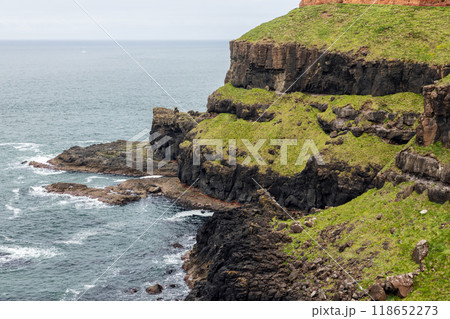 Black basalt cliffs at Giant's Causeway covered in green grass, waves crash against rugged shore 118652273