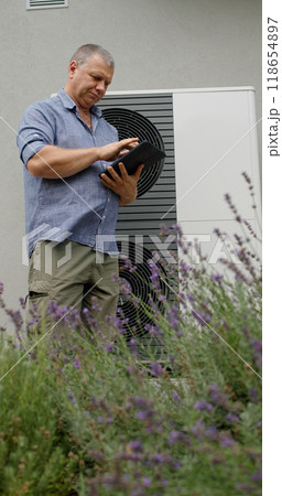 A homeowner checks a heat pump unit's settings on a tablet amidst lavender. A homeowner checks a heat pump unit's settings on a tablet amidst lavender. 118654897