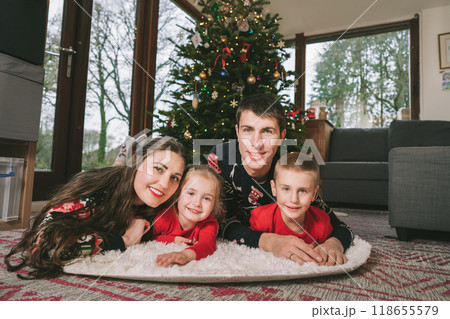 Smiling family portrait in festive sweaters laying under decorated Christmas tree at home during holiday. Happy parents and kids cuddling, feeling excited. Family time together. Happy childhood. 118655579