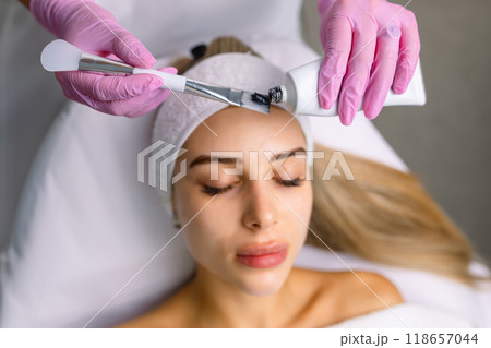 cosmetologist applying black mask on pretty woman face wearing black gloves, gorgeous woman in spa having facial procedures 118657044