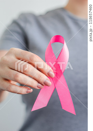 Woman in gray t-shirt holds pink ribbon in her hands. Breast Cancer Awareness Month. Supporting and promoting campaign against cancer. Health care concept. Vertical photo. Woman in gray t-shirt holds pink ribbon in her hands. Breast Cancer Awareness Month. Supporting and promoting campaign against cancer. Health care concept. Vertical photo. 118657460