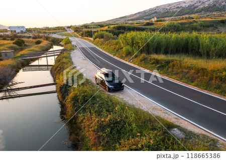 A black vehicle equipped with a roof rack is parked along a serene road, surrounded by greenery 118665386