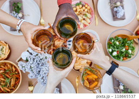 Four diverse hands of friends clinking glasses of mixed drinks over a table with various dishes, symbolizing unity and friendship 118665537