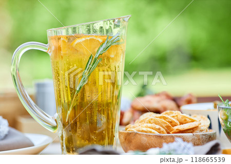 Refreshing citrus-infused beverage in elegant glass pitcher with rosemary sprig, accompanied by snacks on wooden table in bright outdoor setting with blurred background Refreshing citrus-infused beverage in elegant glass pitcher with rosemary sprig, accompanied by snacks on wooden table in bright outdoor setting with blurred background 118665539