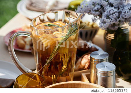 Refreshing iced tea served in clear pitcher adorned with lemon slices and decorative rosemary next to flowers and seasoning containers on outdoor table setup 118665573