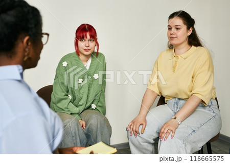 Three individuals engaging in therapy session, including therapist and two women on chairs. One woman has red hair while the other has brown hair, both wearing casual clothes Three individuals engaging in therapy session, including therapist and two women on chairs. One woman has red hair while the other has brown hair, both wearing casual clothes 118665575