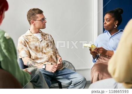 Two colleagues engaging in conversation about project details in comfortable office setting. One holding notebook while other listens attentively wearing casual attire 118665593