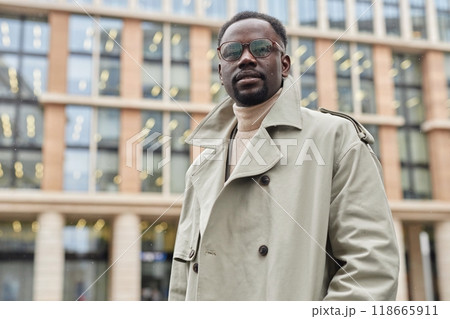 Portrait of African American man standing in urban setting, wearing beige trench coat and sunglasses, looking straight at camera, exuding confidence and sophistication Portrait of African American man standing in urban setting, wearing beige trench coat and sunglasses, looking straight at camera, exuding confidence and sophistication 118665911