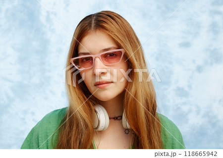 Portrait of young Hispanic woman with long hair wearing pink glasses and white headphones, standing against blue textured background Portrait of young Hispanic woman with long hair wearing pink glasses and white headphones, standing against blue textured background 118665942