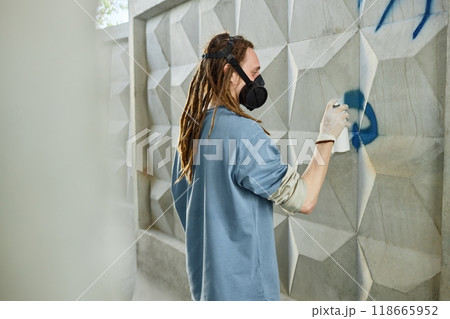 Artist is standing near geometric wall while creating abstract street art using spray paint. Person is wearing protective gear and face mask for safety 118665952