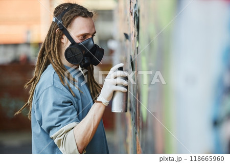 Artist wearing protective mask and gloves concentrating on creating colorful mural on city wall, adding intricate details with spray paint can, showcasing urban graffiti culture 118665960