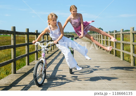 Two girls are riding bicycles on a wooden bridge. One girl is mid-air, playfully leaping off her bike, while the other concentrates on riding. The bright day highlights their adventurous spirits Two girls are riding bicycles on a wooden bridge. One girl is mid-air, playfully leaping off her bike, while the other concentrates on riding. The bright day highlights their adventurous spirits 118666135