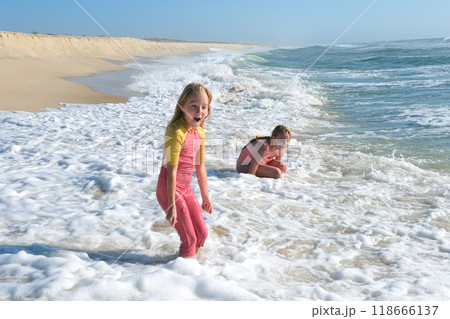 Two young girls are playing in the ocean waves on a sandy beach, laughing and splashing in the foamy water. The sun is shining brightly, creating a fun and carefree summer scene 118666137