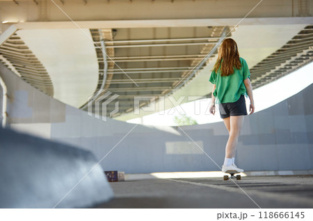 Young person skating under urban bridge during sunny day. Skater wearing casual green shirt and shorts, moving away from camera 118666145