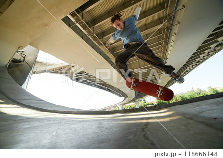 Young male skateboarder performing mid-air trick under urban bridge with concrete structures in background capturing dynamic movement and urban setting 118666148