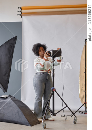 Vertical full length portrait of Black female photographer setting up lighting equipment preparing for creative project in photo studio 118666214