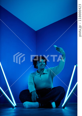 Full length portrait of African American woman taking selfie photo with smartphone while sitting on floor in room with blue neon lights Full length portrait of African American woman taking selfie photo with smartphone while sitting on floor in room with blue neon lights 118666423