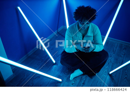 Full length portrait of African American woman using smartphone while sitting on floor in tiny room with blue neon light strips Full length portrait of African American woman using smartphone while sitting on floor in tiny room with blue neon light strips 118666424