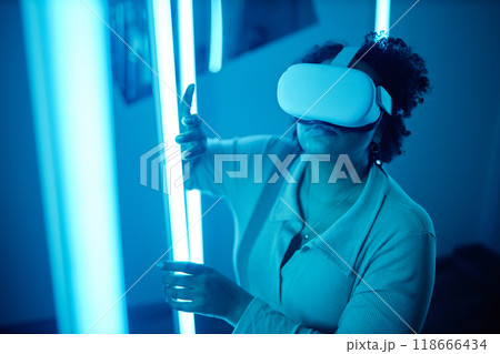 Portrait of African American woman wearing VR headset in modern art show interacting with blue neon lights enjoying multisensory experience 118666434