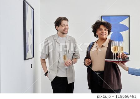 Waist up portrait of two smiling people taking champagne glasses while visiting grand opening ceremony of modern art museum, copy space 118666463