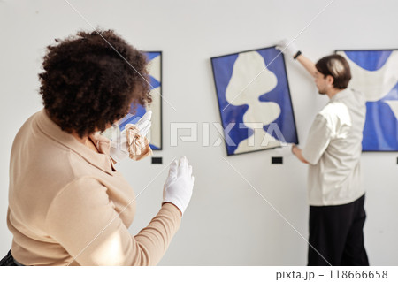 Back view of two art gallery workers hanging paintings on wall and giving directions while planning exhibition copy space Back view of two art gallery workers hanging paintings on wall and giving directions while planning exhibition copy space 118666658