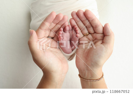 The palms of the parents. A father and mother hold the feet of a newborn child in a white blanket on a white background.. The feet of a newborn in the hands of parents. Photo of foot, heels and toes 118667052