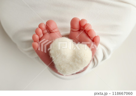 The tiny foot of a newborn baby. Soft feet of a new born in a white blanket. Close up of toes, heels and feet of a newborn. Knitted white heart in the legs of a baby. Macro photography.  118667060