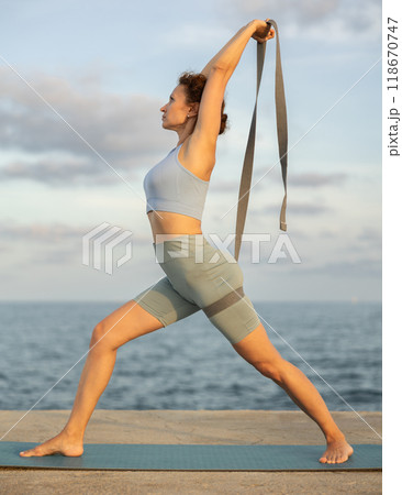 Yoga practice on embankment, female athlete fulfil carry out warrior pose with belt 118670747