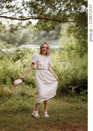 Woman standing in park holding straw hat. Woman standing in park holding straw hat. 118671417