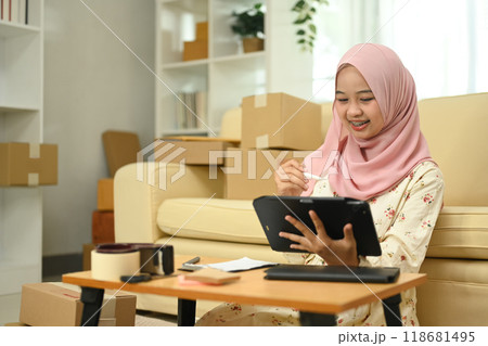 Smiling Asian muslim woman using digital tablet for receiving online purchase order. Small business concept Smiling Asian muslim woman using digital tablet for receiving online purchase order. Small business concept 118681495