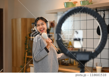 Cheerful young woman holding a wooden spoon, live streaming or recording a video for social media in the kitchen 118681510