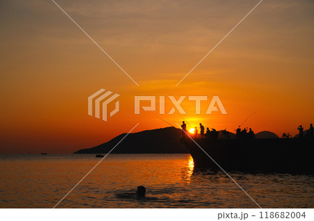 Silhouettes of fishermen with fishing rods fishing on pier in sea in evening at sunset in summer Silhouettes of fishermen with fishing rods fishing on pier in sea in evening at sunset in summer 118682004