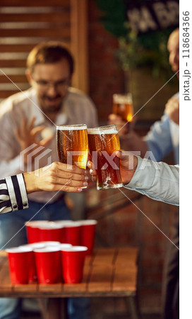 Focused on toast, while blurred figures behind cheer and engage, perfect scene of friends celebrating their team's victory with cold drinks. Focused on toast, while blurred figures behind cheer and engage, perfect scene of friends celebrating their team's victory with cold drinks. 118684366