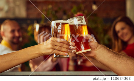 Close-up of two raised glasses filled with amber beer, with blurry background action suggesting lively sports crowd enjoying game together. 118684368