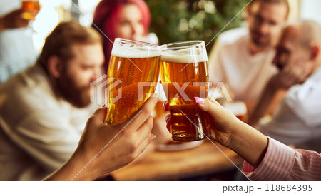 Close-up of fans clinking glasses, beer foaming at top. Behind on blurred background, group adds energy to festive scene. 118684395