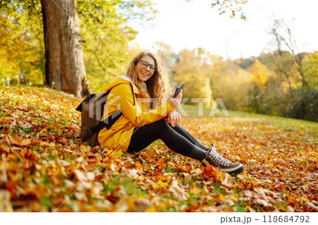 A smiling woman sits outdoors on the lawn among yellow autumn leaves with a phone in her hands. A smiling woman sits outdoors on the lawn among yellow autumn leaves with a phone in her hands. 118684792