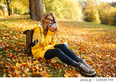 Woman traveler in yellow coat sits in clearing among yellow leaves and drinks hot drink from thermos Woman traveler in yellow coat sits in clearing among yellow leaves and drinks hot drink from thermos 118684794