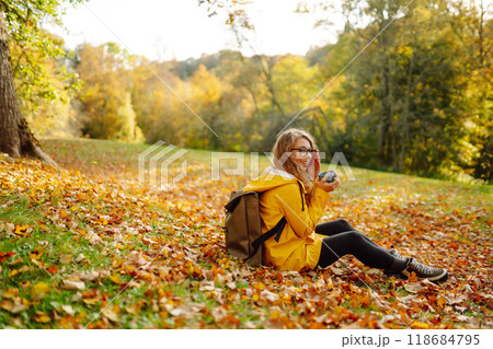 Woman traveler in yellow coat sits in clearing among yellow leaves and drinks hot drink from thermos Woman traveler in yellow coat sits in clearing among yellow leaves and drinks hot drink from thermos 118684795